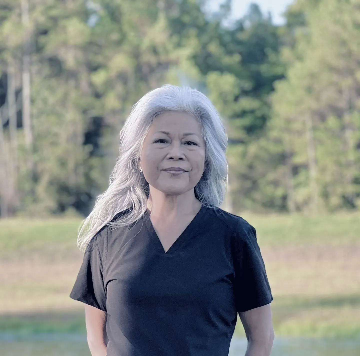 A woman with long white hair standing in front of trees.